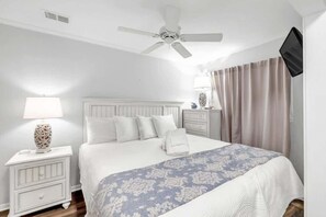 Quarto com cama, mesas de cabeceira, ventoinha no teto, televisão e cortinas.
