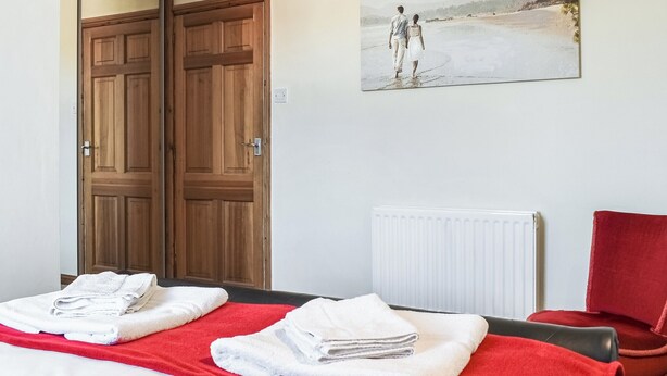 A hotel room with a bed, a red blanket, white towels, a red chair, a wooden door, and a painting of a beach scene. - 2 bedroom accommodation in Whitby (Whitby)