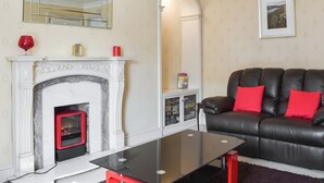 A living room with a fireplace, a black leather sofa with red cushions, a glass coffee table, and a framed picture on the wall. - 2 bedroom accommodation in Whitby (Whitby)