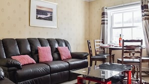 A living room with a black leather sofa, a glass coffee table, and a dining table with chairs. - 2 bedroom accommodation in Whitby (Whitby)