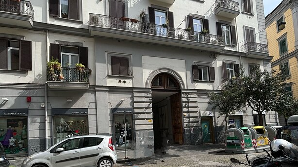 Ein mehrgeschossiges Gebäude mit Eingang im Erdgeschoss, ein Auto vor der Fassade und ein Roller rechts daneben.