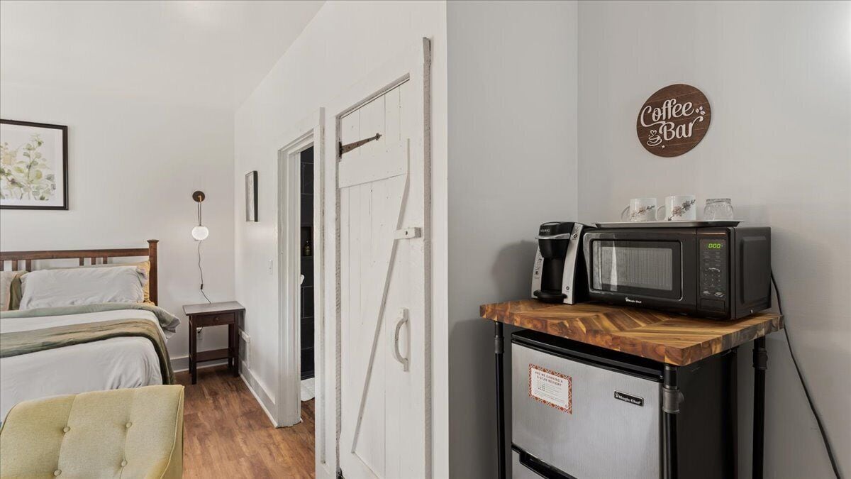 A bedroom with a bed, bedside table, lamp, and a kitchenette with a microwave and coffee maker.