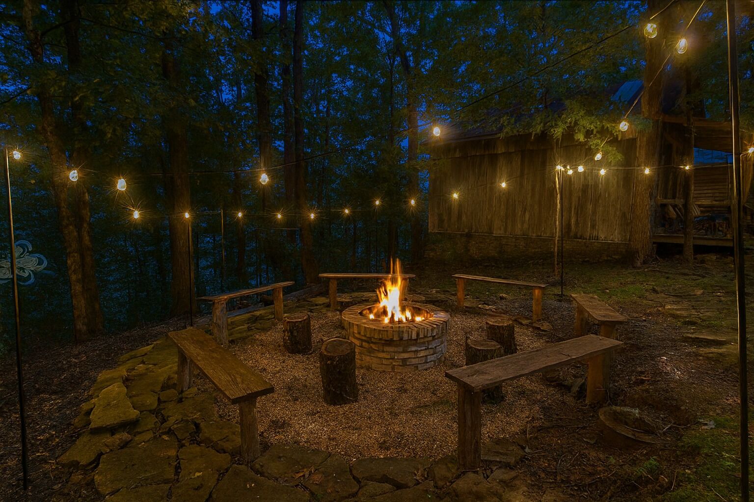 A campfire surrounded by wooden benches and a stone-paved area, with string lights overhead.