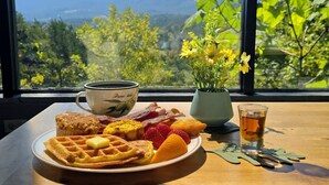 Una mesa de desayuno con waffles, fresas, una taza de café, un florero y una taza de té, con vista a la montaña a través de una ventana.