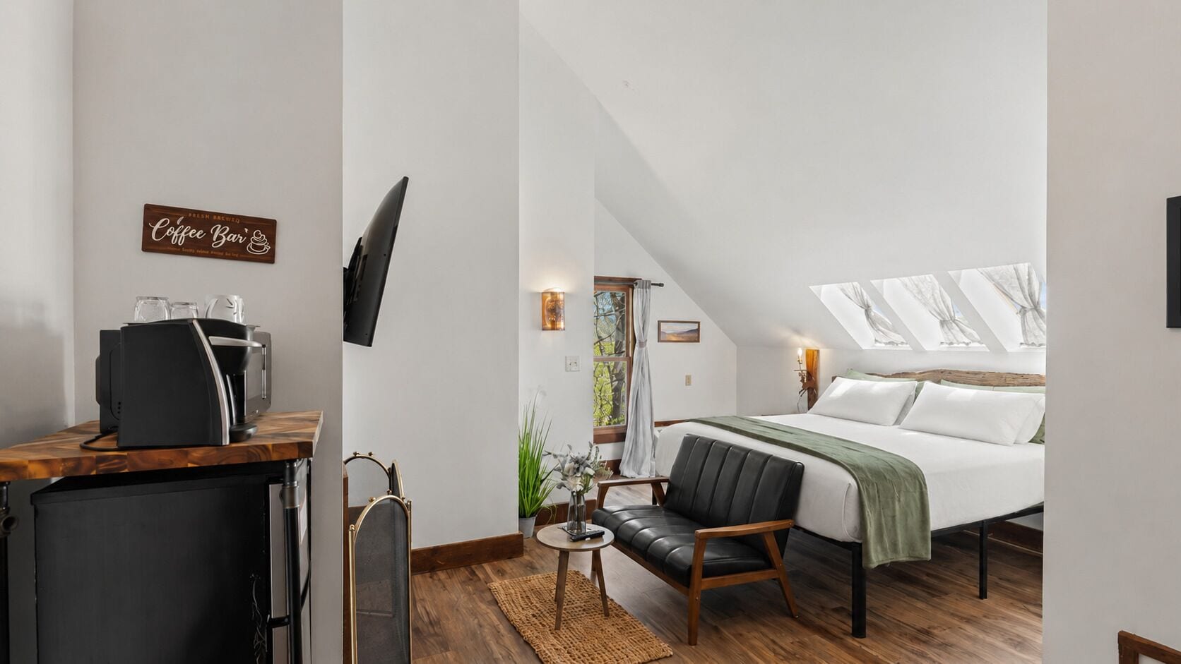 A modern bedroom with a bed, a desk with a coffee machine, a TV, and a view of the outdoors through skylights.