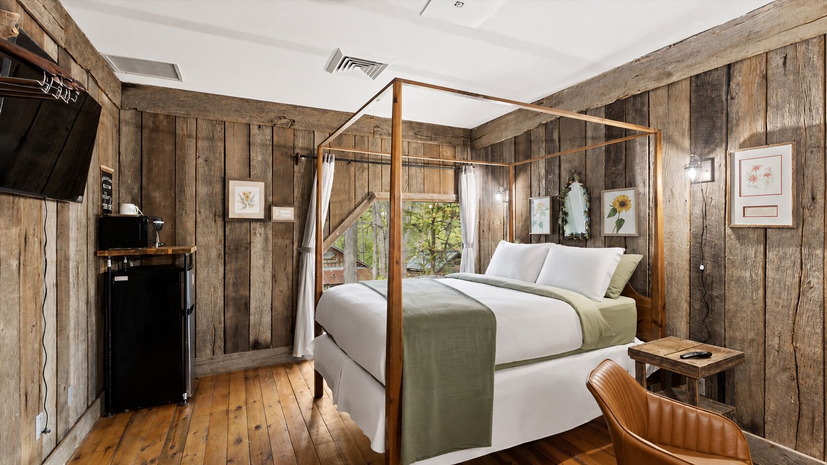 Un letto a baldacchino con struttura in legno, un comodino, una sedia e un frigorifero in una stanza rustica con pareti in legno e vista sul verde circostante.