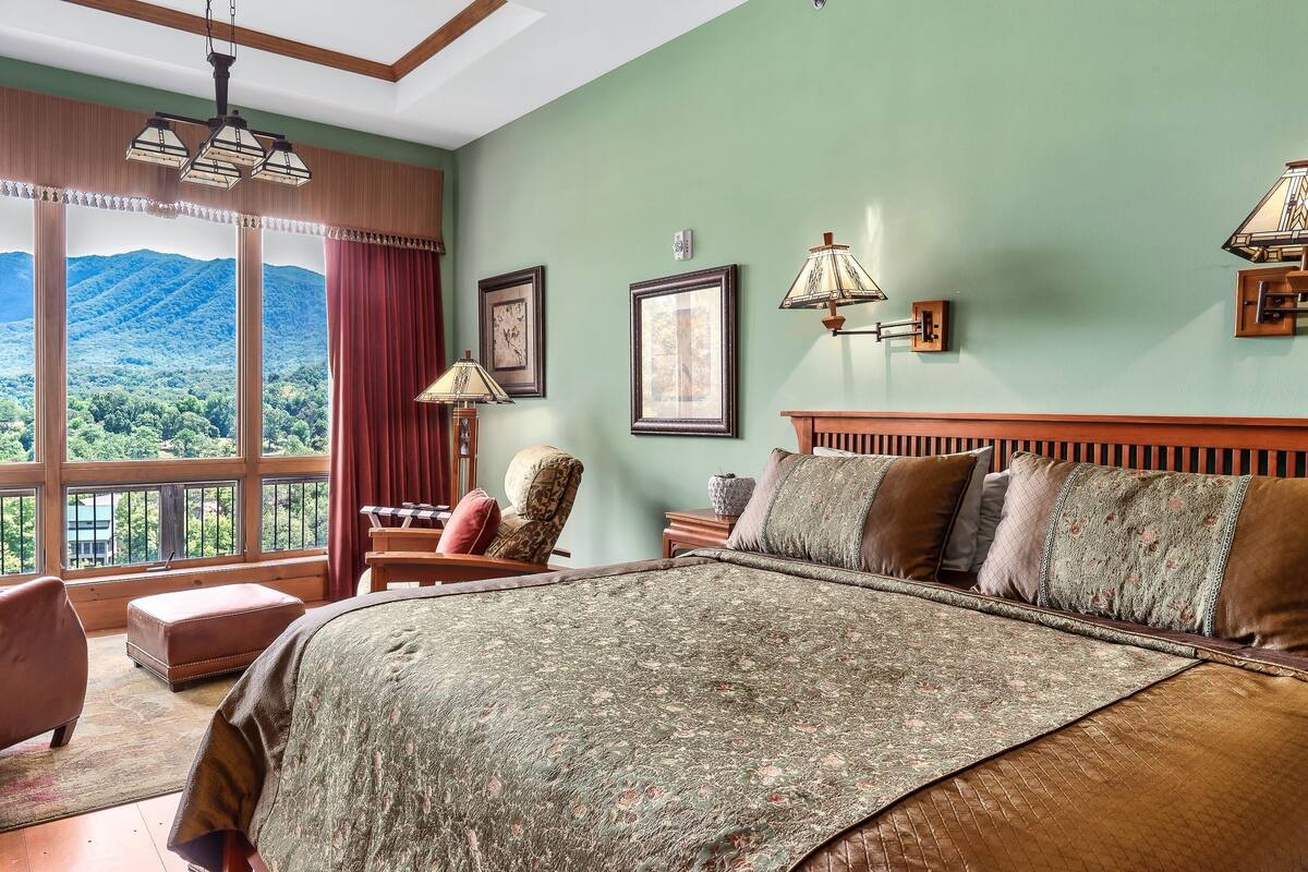 A bedroom with a large bed, a chair, a nightstand, and a view of mountains through large windows.