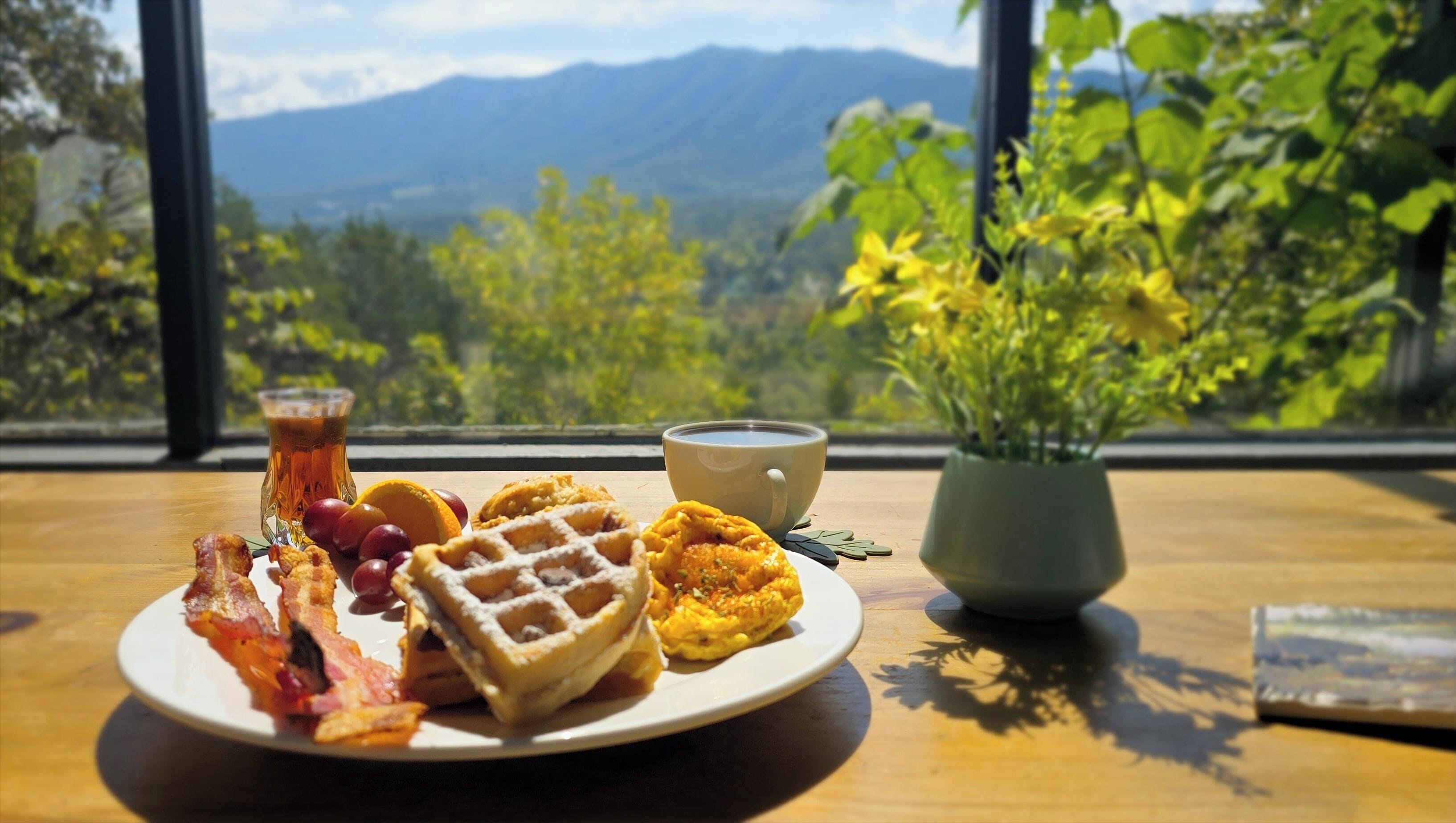 A breakfast plate with waffles, bacon, and fruit, a cup of coffee, and a vase of flowers on a table by a window with a mountain view.
