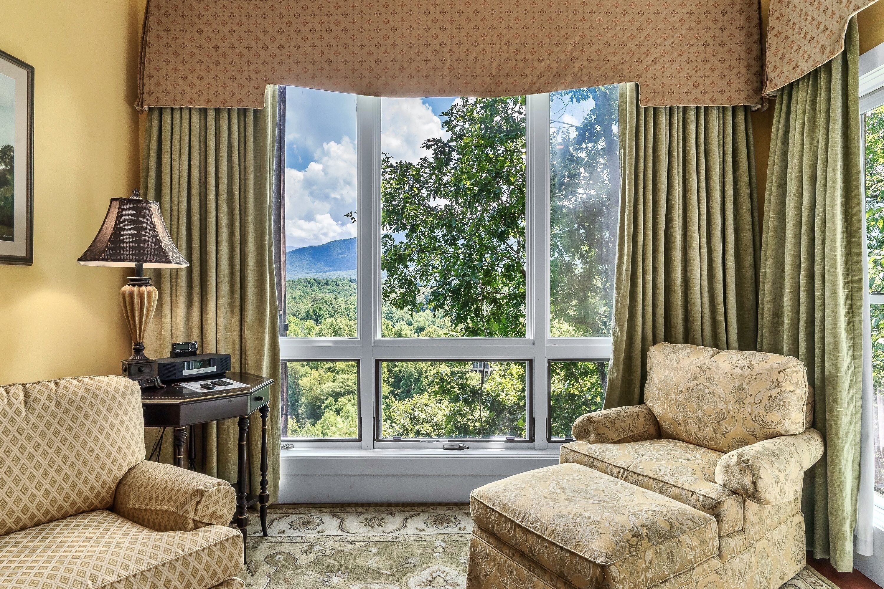 A room with a large window, a desk with a lamp, and two patterned armchairs.
