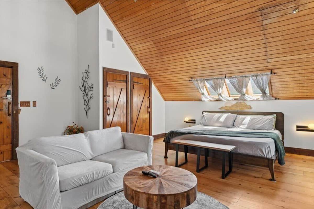 A bedroom with a bed, a sofa, a wooden coffee table, and a large window with curtains.