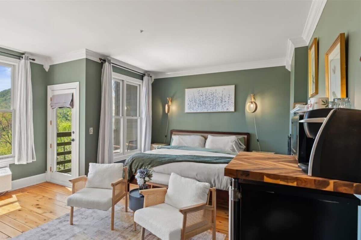 A bedroom with a bed, two chairs, a desk, a heater, and a window with curtains.