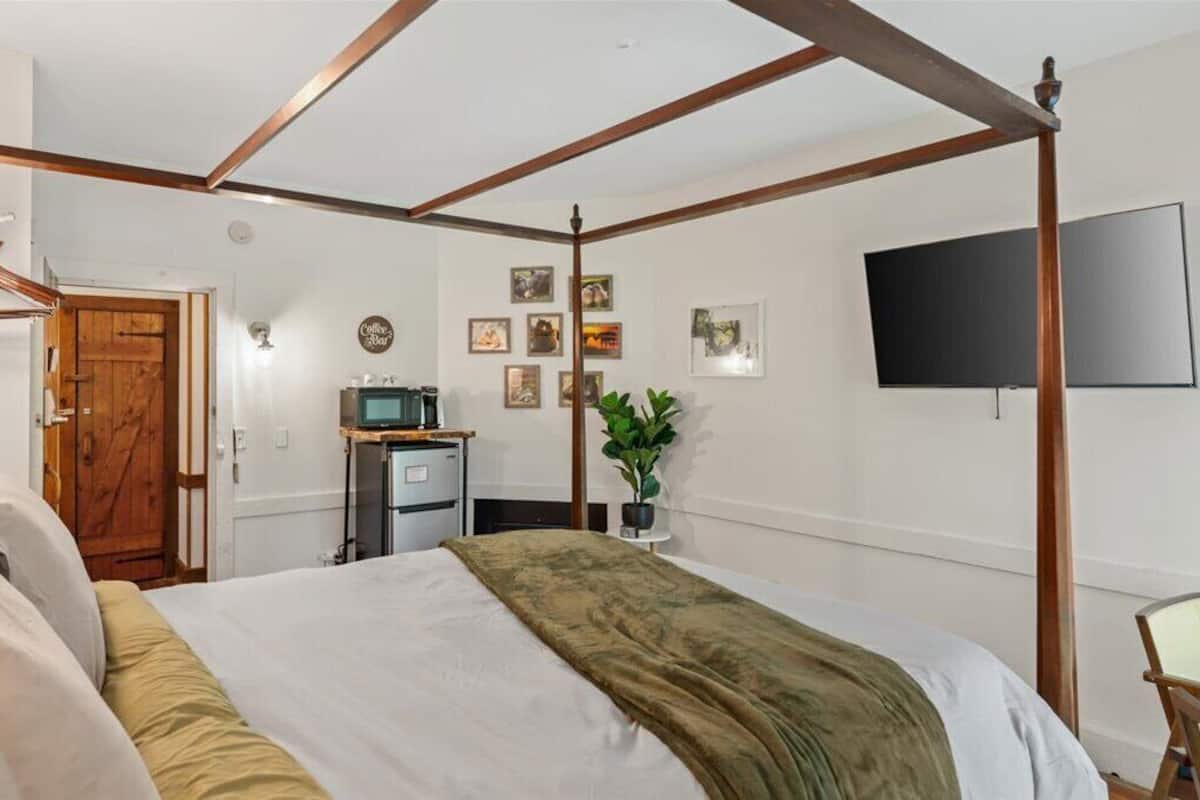 A bedroom with a four-poster bed, a television mounted on the wall, a small desk with a computer, and a potted plant.