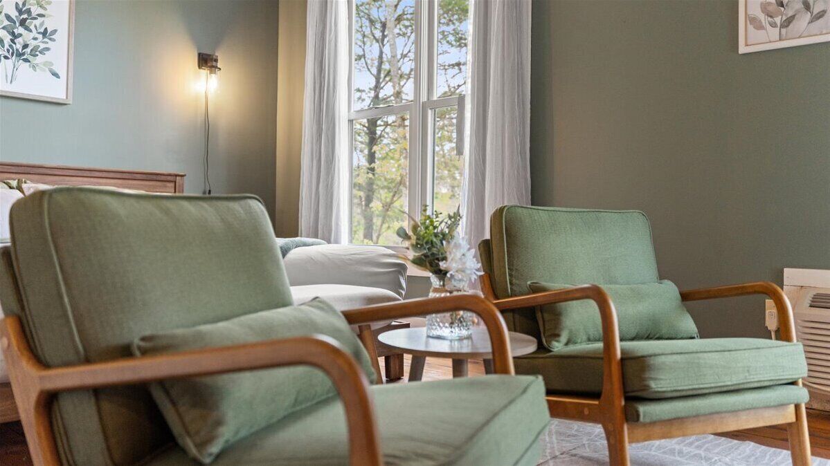 Une pièce avec deux fauteuils verts, une petite table ronde et un lit à l’arrière-plan.