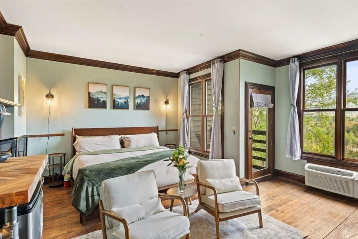 A bedroom with a bed, two chairs, a table, a radiator, and large windows with curtains.