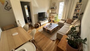 Un salon confortable avec un canapé, une table basse et un téléviseur. On y trouve également un coin repas avec des chaises et une table. Une bibliothèque et une plante sont également visibles.