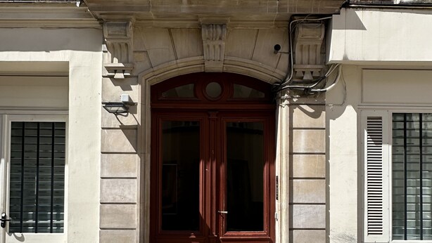 Une façade d’immeuble avec une entrée centrale, ornée d’une double porte en bois, flanquée de deux fenêtres avec volets, et un sol pavé de pierres.