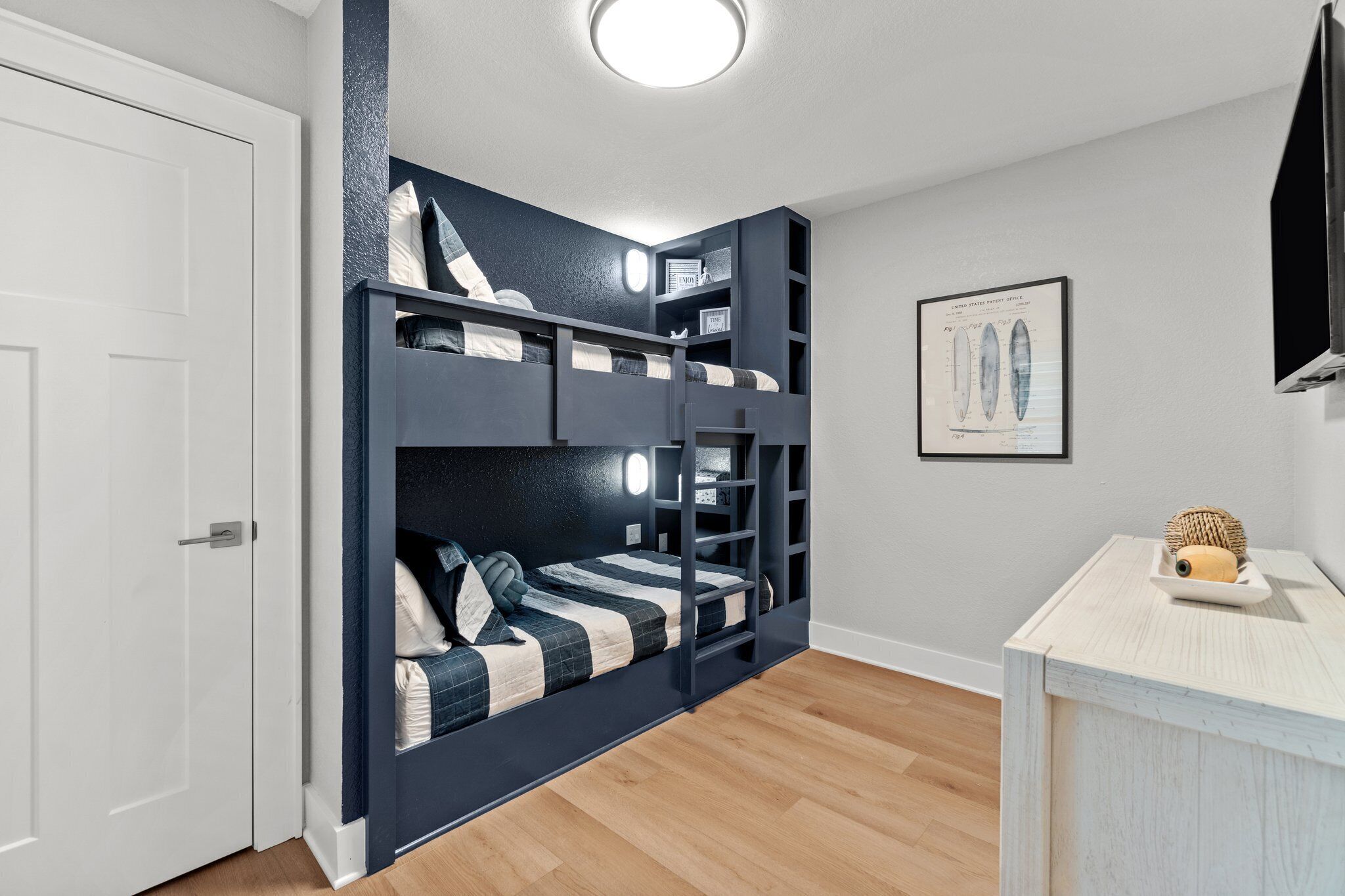 A room with a bunk bed, a white door, a wooden floor, a small table with a basket, and a framed picture on the wall.