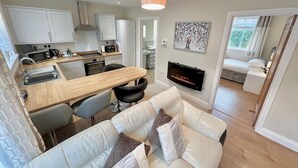 A modern kitchen with white cabinets, a wooden dining table, and black chairs. There is a fireplace with a TV mounted above it, and a white sofa with cushions in the living area. - Modern Cosy home, Limeslade Bay - Pass the Keys (The Mumbles)
