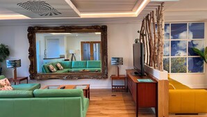 Ein Wohnzimmer mit einem grünen Sofa, einem gelben Hocker, einem großen Spiegel und einem Holztisch.