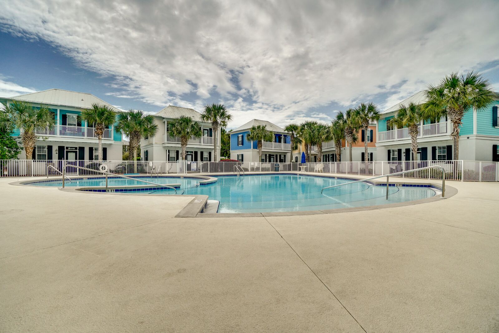 A swimming pool area with multiple pools, surrounded by a white fence, and buildings with balconies and palm trees in the background.
