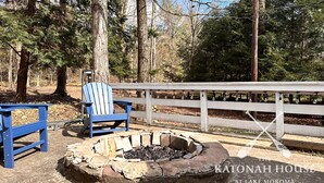 A fire pit with a blue Adirondack chair and a white fence in a wooded area.