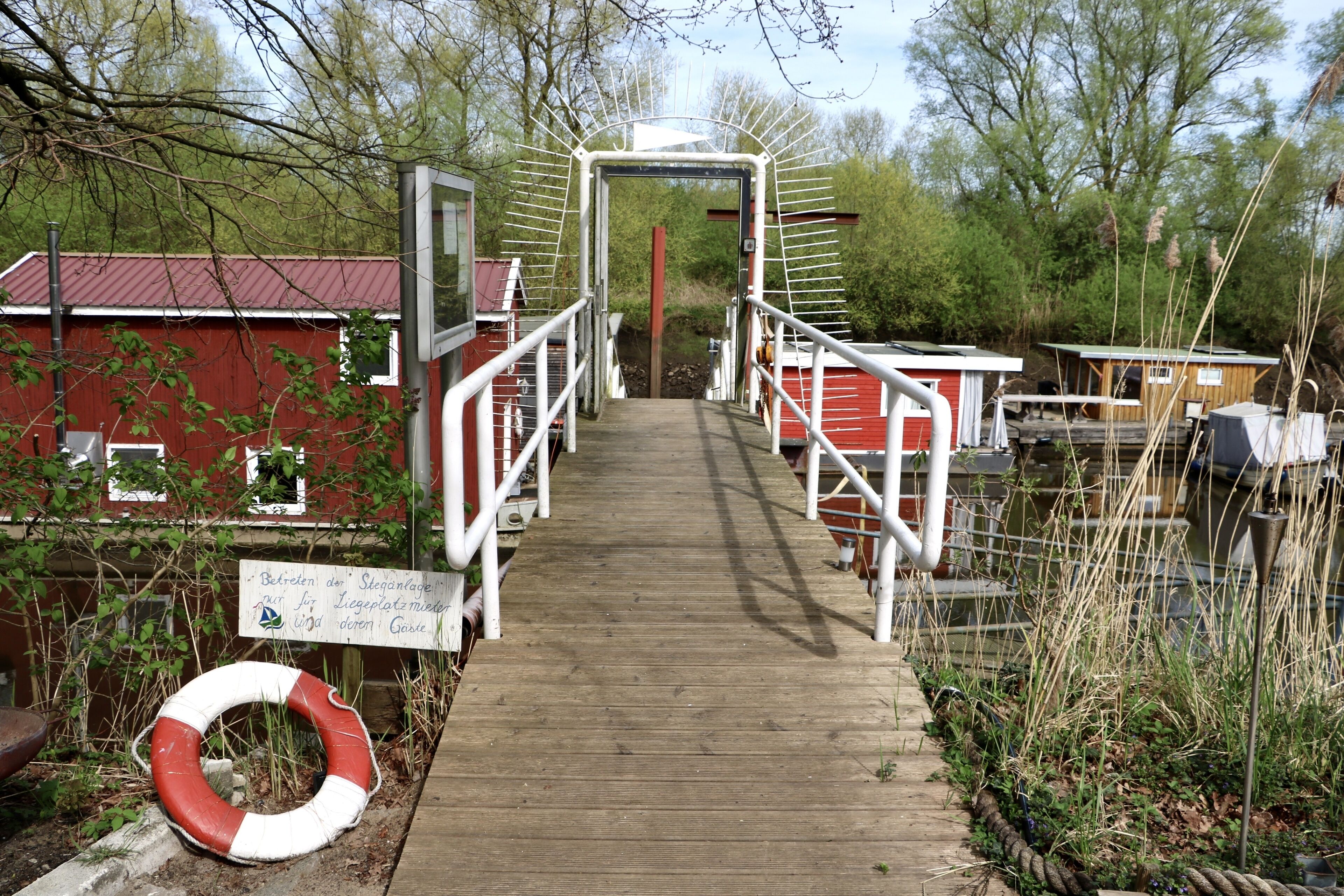 Un chemin en bois mène à un bâtiment doté d’un toit rouge, entouré de verdure et surmonté d’une bouée de sauvetage posée au sol.