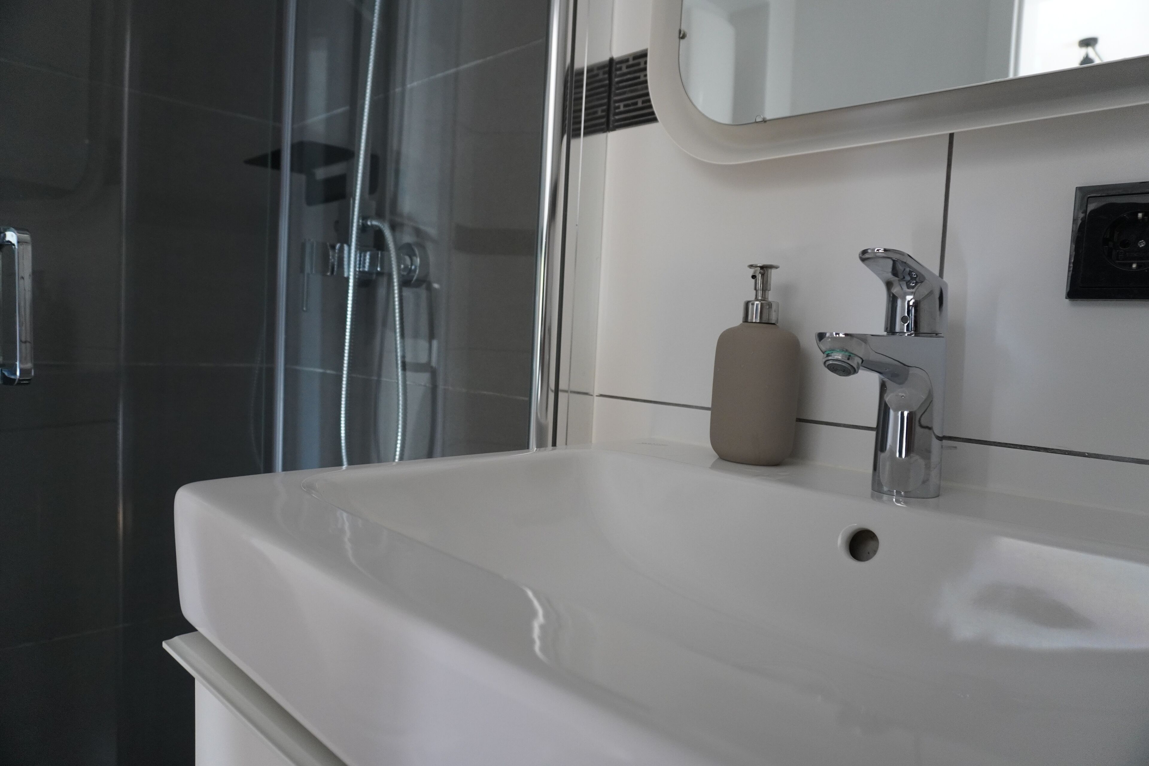 Baño con lavamanos blanco, espejo, dispensador de jabón y zona de ducha.