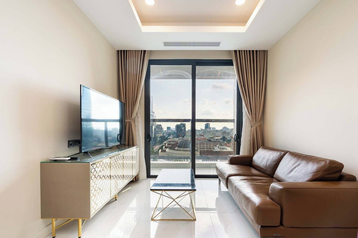 A modern living room with a sofa, a TV on a cabinet, and a city view through large windows.