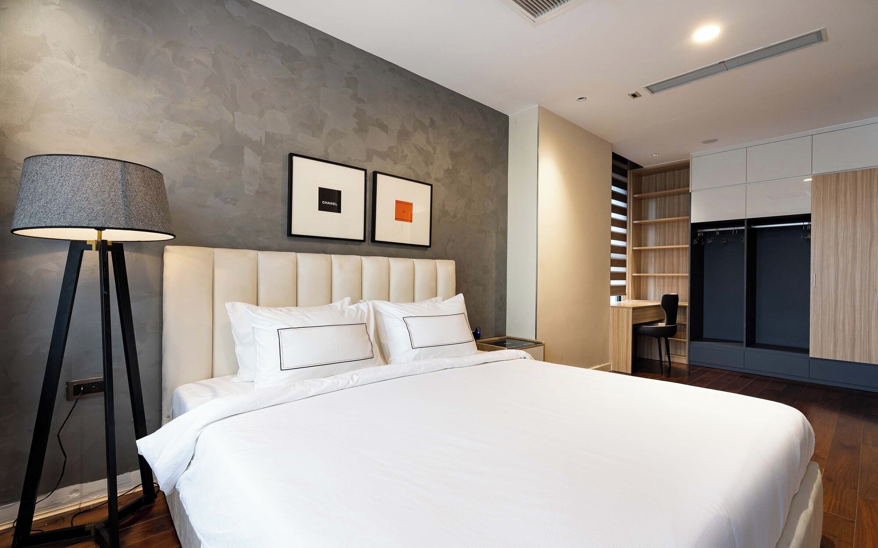 A modern hotel room with a large bed, a standing floor lamp, a wardrobe, and a desk with a chair.