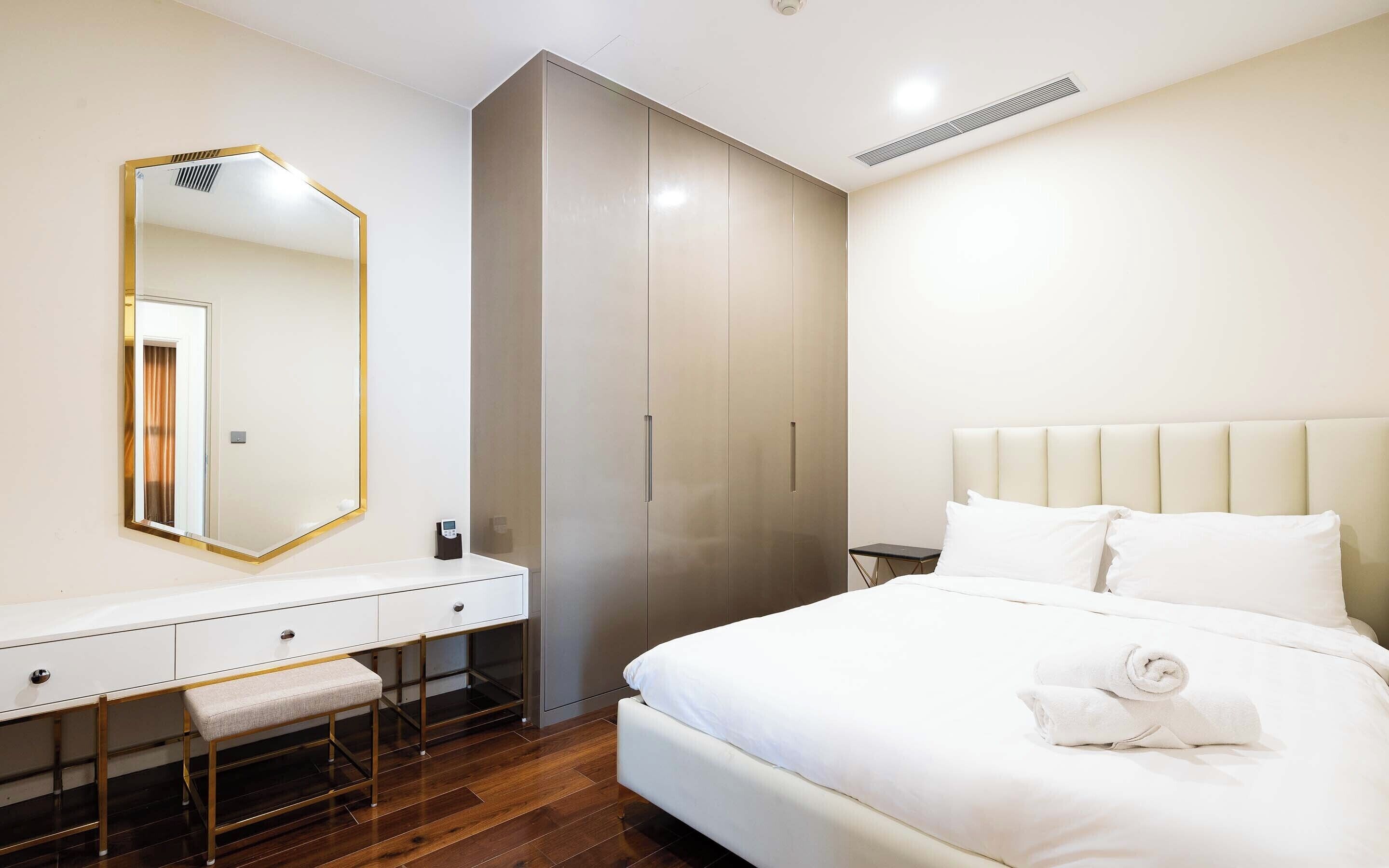 A modern hotel room with a large bed, a vanity with a mirror, and a wardrobe.