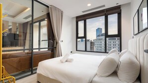 Ein modernes Hotelzimmer mit einem großen Bett, Blick auf die Stadt durch raumhohe Fenster, einem Fernseher und einem Balkon mit gelber Brüstung.
