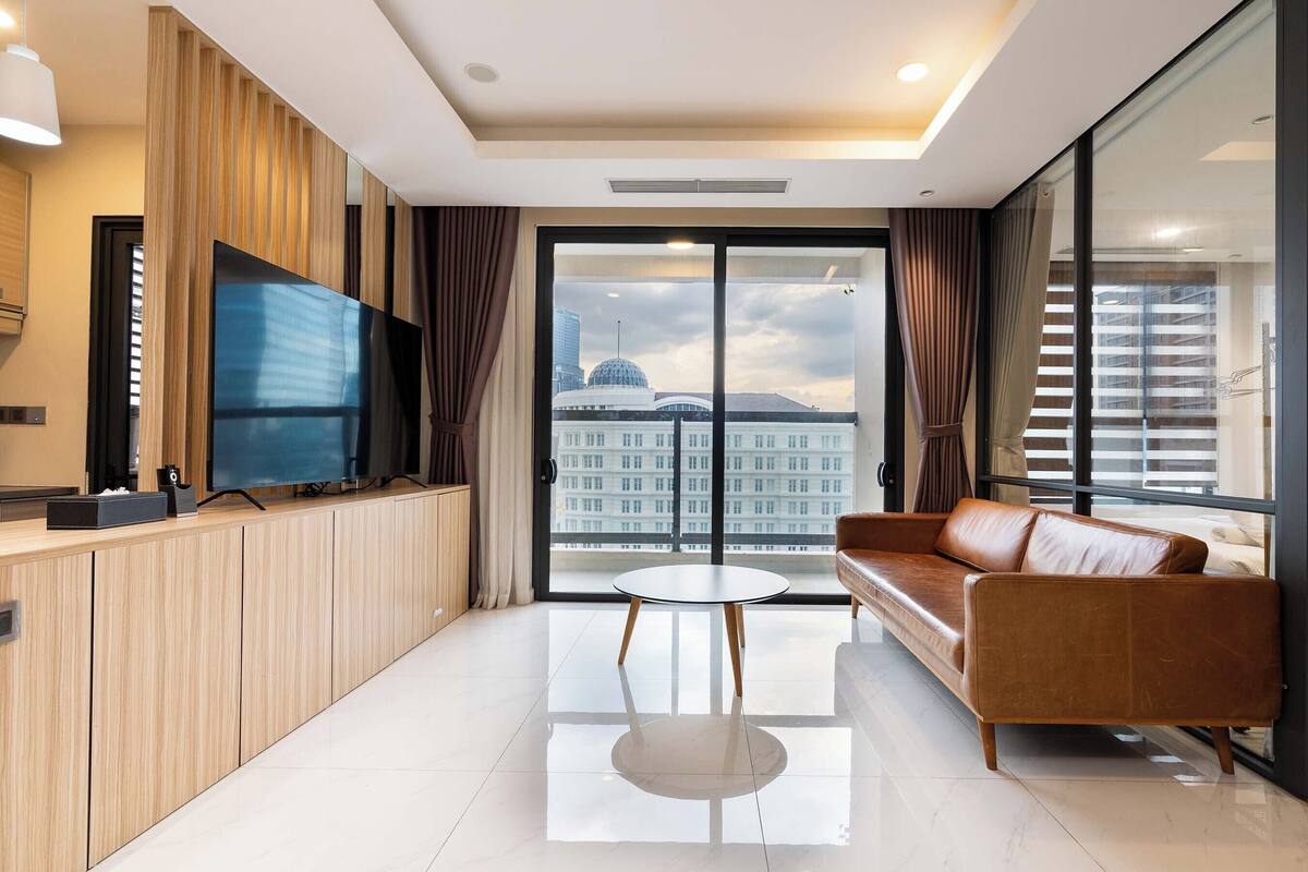 A modern living room with a large TV, a leather sofa, a small round table, and a view of a cityscape through large windows.
