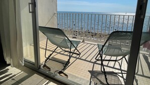 Ein Balkon mit zwei Stühlen und einem Paar Schuhen auf dem Boden, mit Blick auf einen Strand mit Menschen und einem Segelboot.