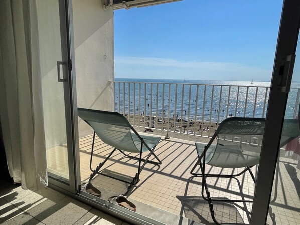A balcony with two chairs and a pair of shoes on the floor, overlooking a beach with people and a sailboat.