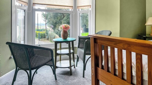 A corner room with a bay window, a small table with a vase, two chairs, and a crib. - Escape to our pet-friendly haven nestled in the heart of Carmarthen. (Carmarthen)