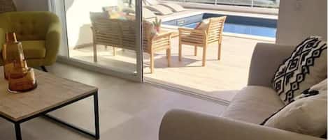 A living room with a sofa, a coffee table, and a yellow armchair. There is a sliding glass door leading to a balcony with a pool and lounge chairs.