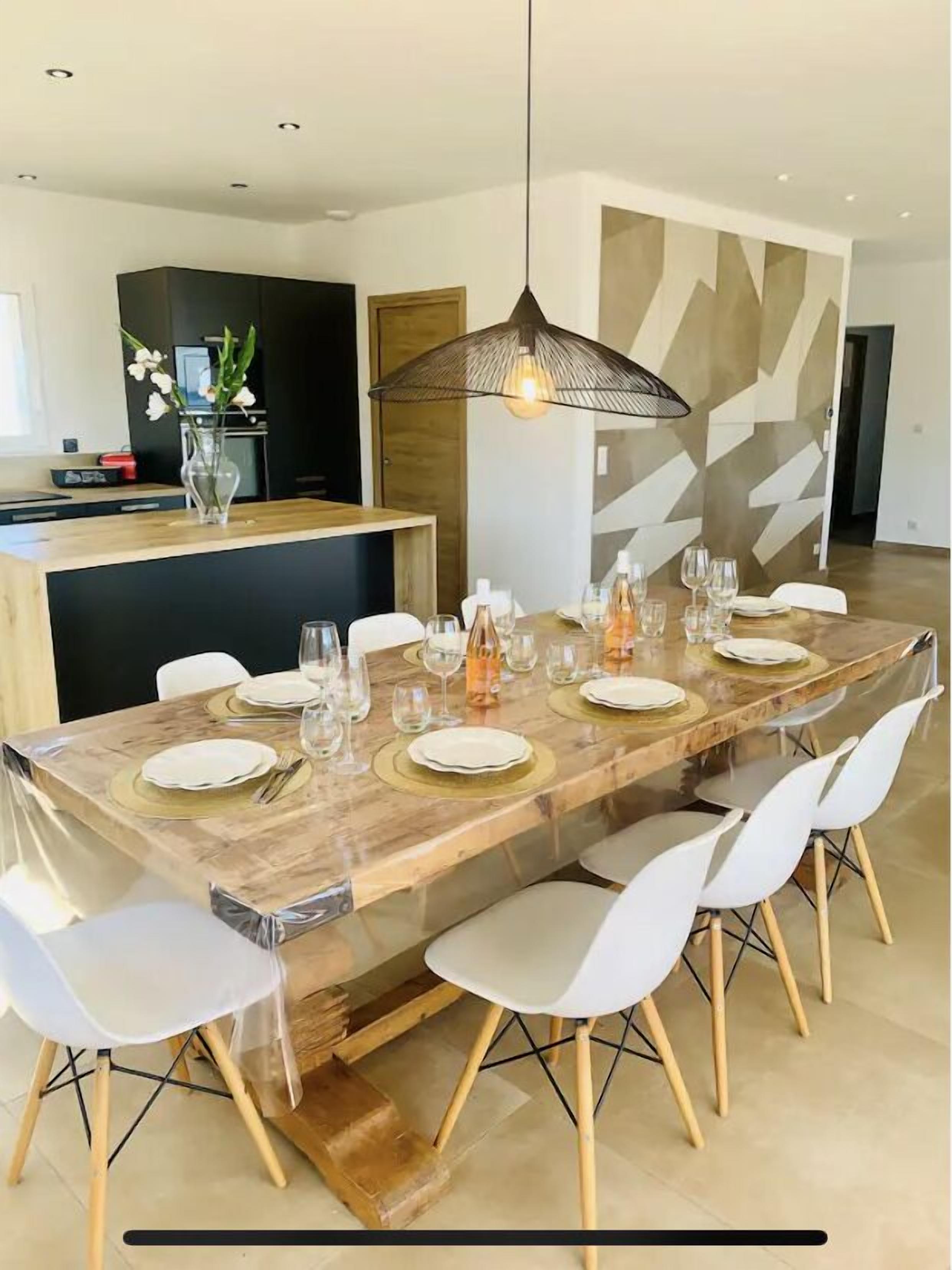 Un comedor moderno con una larga mesa de madera dispuesta para seis personas, sillas blancas y una lámpara colgante.