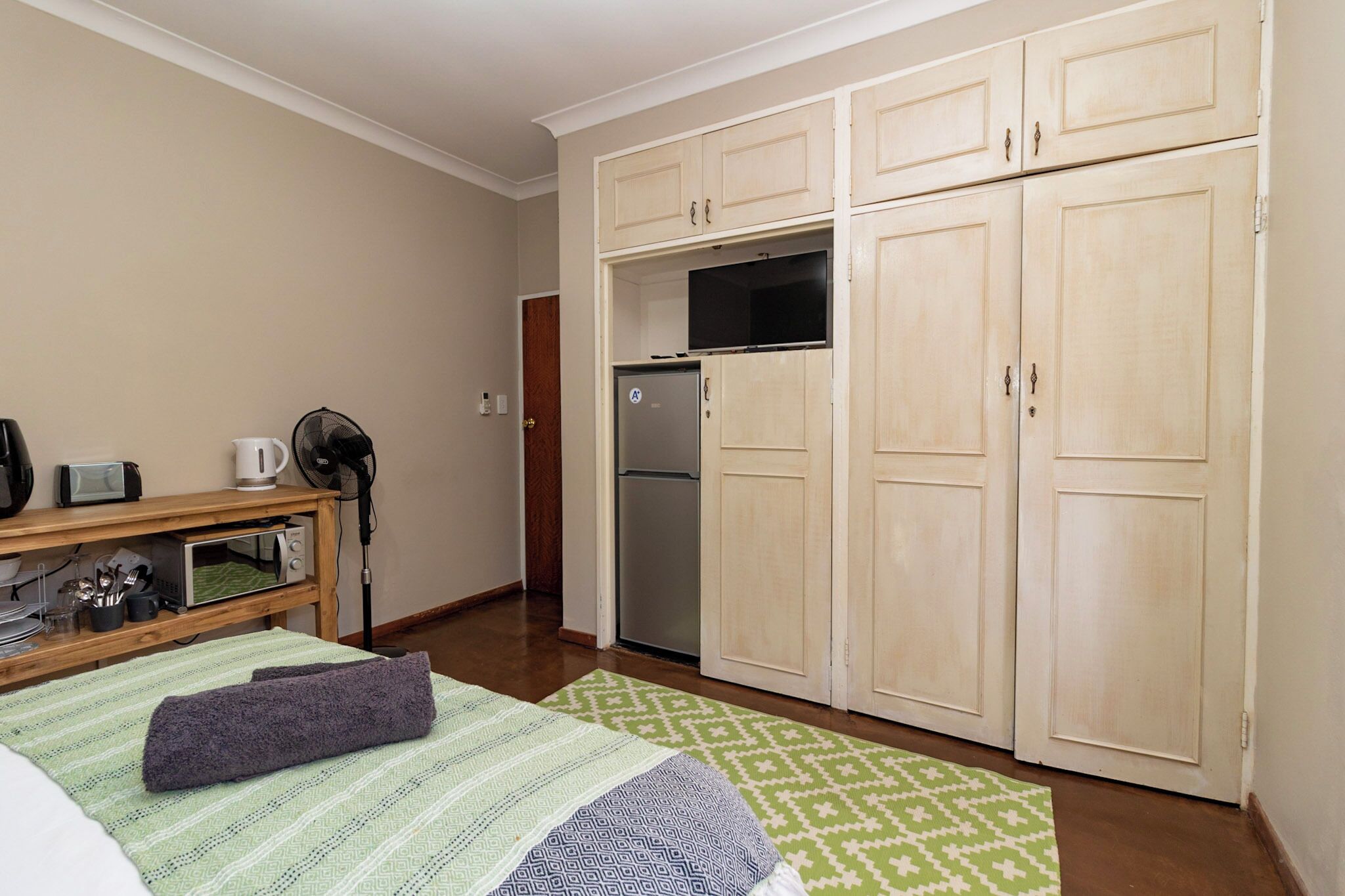 A compact room with a bed, a small table with a kettle, a fan, a microwave, and a refrigerator.