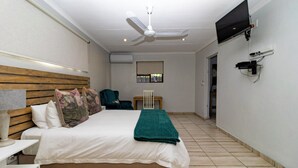 Une chambre à coucher avec un lit, un ventilateur de plafond, un téléviseur fixé au mur et une fenêtre avec des stores.
