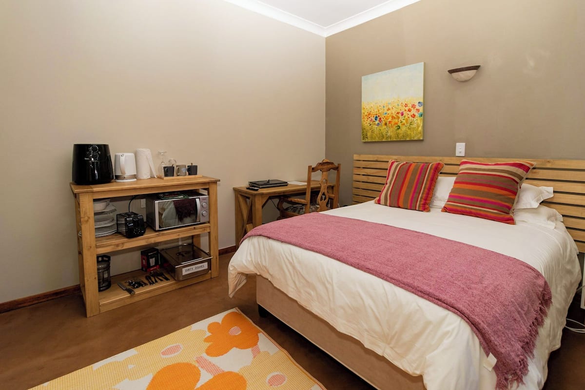 Een slaapkamer met een bed, een houten nachtkastje, een plank met diverse spullen en een schilderij aan de muur.