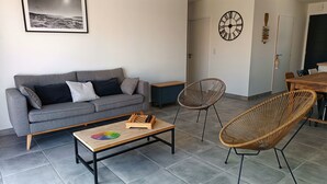 Uma sala de estar com um sofá cinzento, uma mesa de centro de madeira, duas cadeiras de vime, uma mesa de jantar com cadeiras, um relógio de parede e um quadro emoldurado.