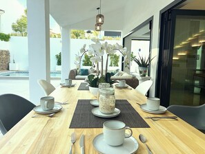 A dining area with a wooden table set for two, white chairs, and a view of the outdoors. - Moderna Villa con Piscina, a 30 Metros de la Playa, en la Mejor Zona de Marbella (Marbella)