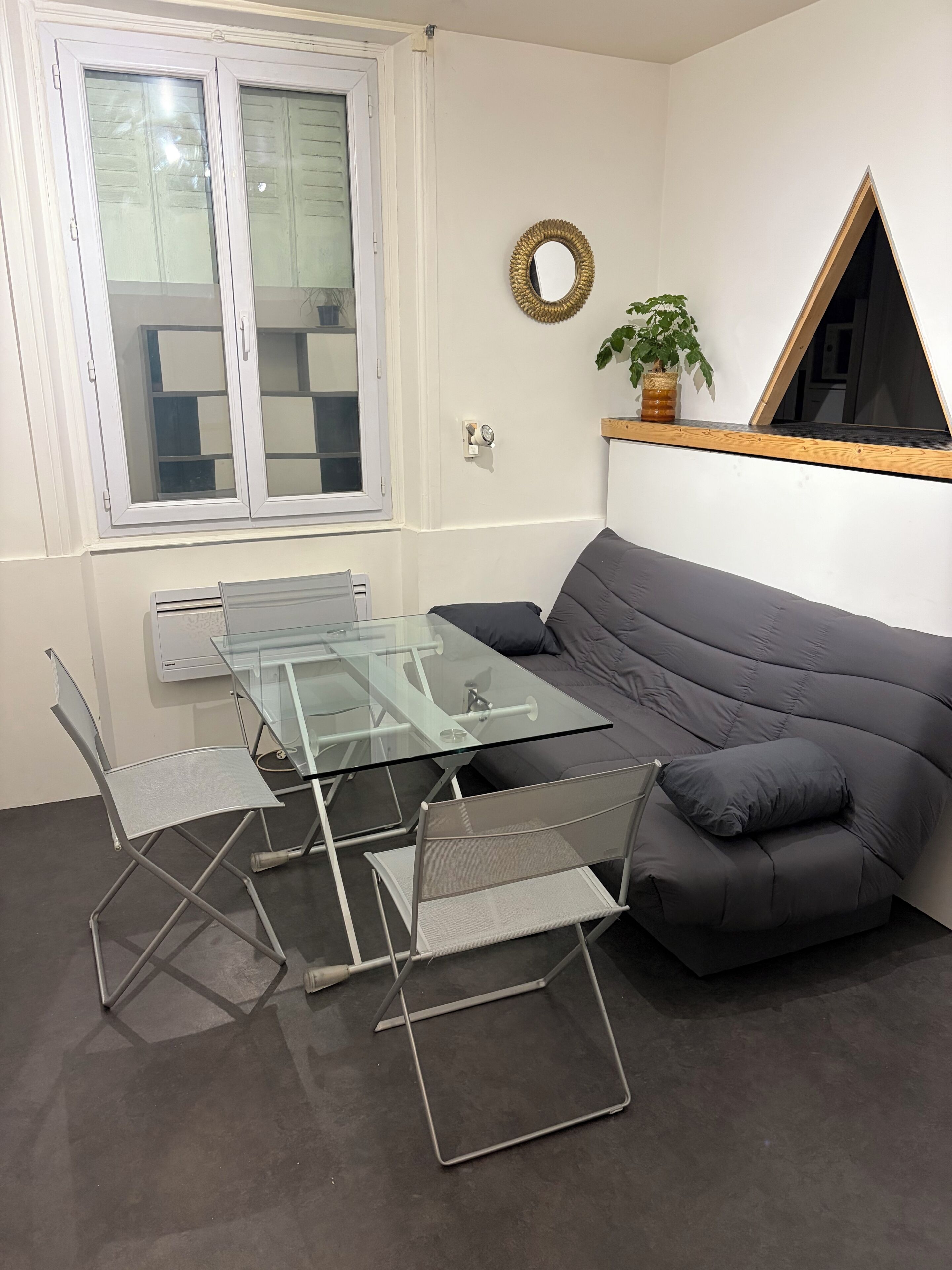 A modern living room with a glass-top coffee table, metal chairs, a grey sofa, a window with blinds, a wall-mounted mirror, and a small plant.
