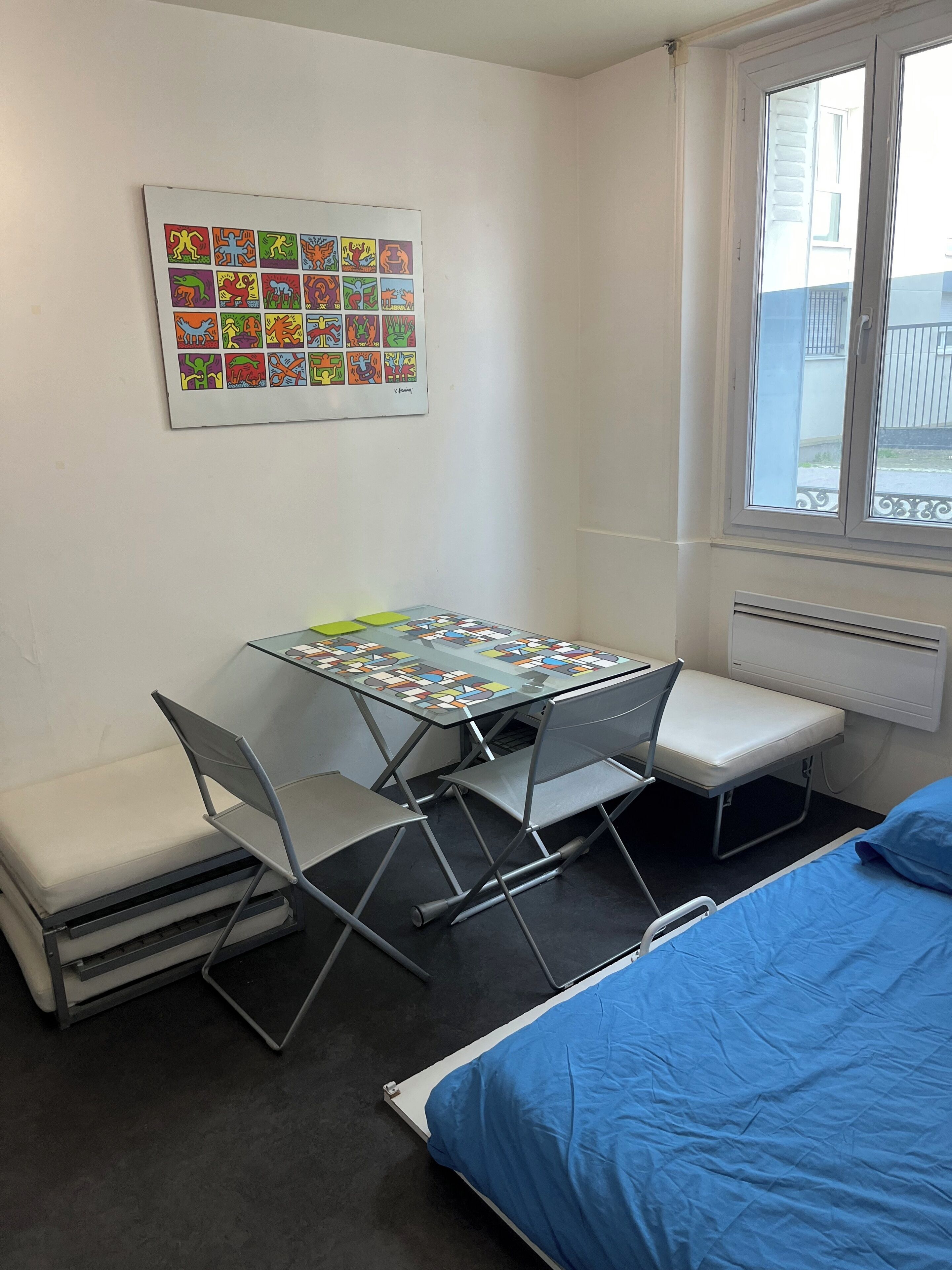 A compact room with a foldable table, two chairs, a bench, a bed with blue bedding, and a window with a view of the outside.
