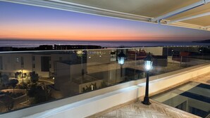 A balcony with a view of the sunset over the ocean, a glass railing, and a wooden floor.