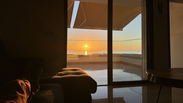 A room with a sliding glass door leading to a balcony with a view of the sunset over the ocean.