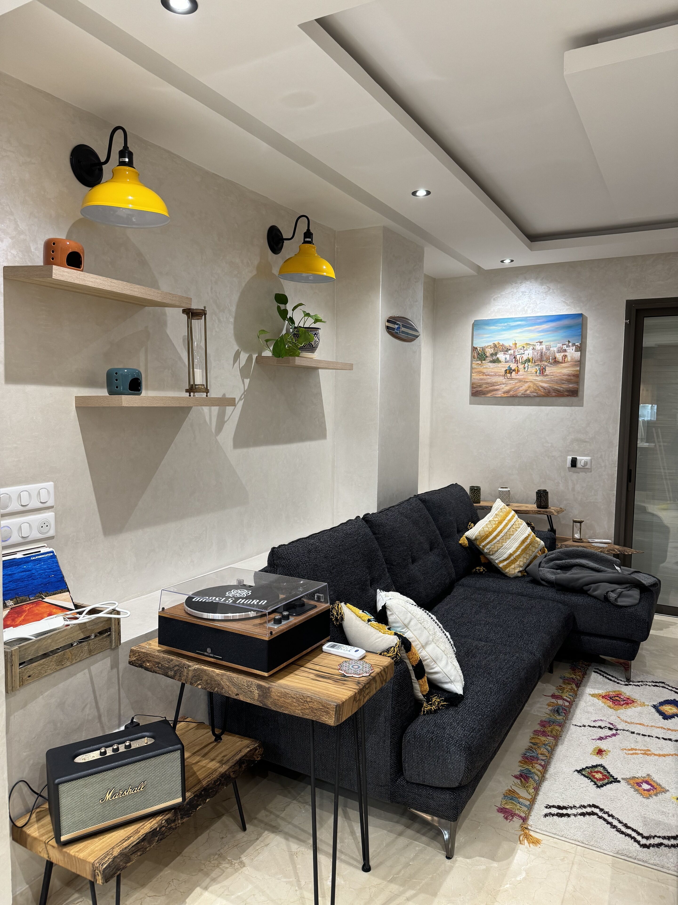 A modern living room with a dark grey sofa, a wooden coffee table with a turntable and Marshall amplifier, a yellow pendant light, and a painting on the wall.