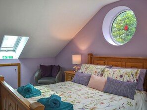 Um quarto com teto inclinado, uma cama com roupa de cama florida, uma janela redonda com uma flor vermelha, uma cadeira e uma mesa de cabeceira com uma luminária.