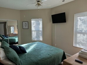Ein Schlafzimmer mit einem Bett, einem Deckenventilator, einem an der Wand befestigten Fernseher und einem Fenster mit Jalousien.