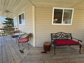 Eine überdachte Veranda mit Holzboden, einem runden Tisch mit zwei Stühlen, einer Bank mit rotem Kissen und einem Fenster mit Jalousien.